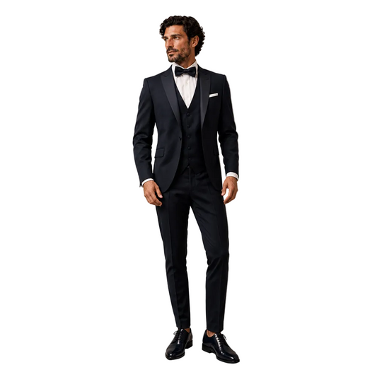 A man standing against a neutral textured studio background modeling a Digel men's ceremony tuxedo in black. The formal evening suit features a tailored dinner jacket with black satin peak lapels, a matching black waistcoat, and slim trousers, accessorized with a white tuxedo shirt, a dark bow tie, and polished black dress shoes