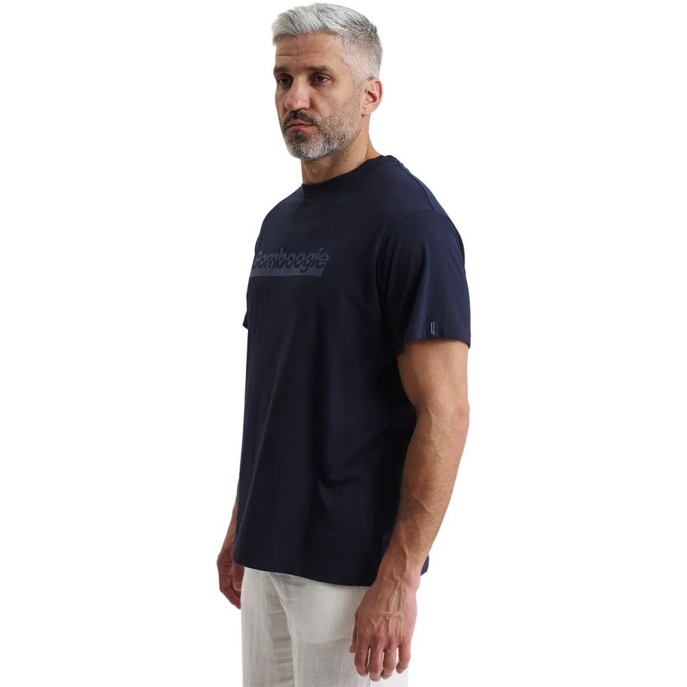 A man modeling a Bomboogie men's navy blue t-shirt against a plain white background. The short-sleeve crewneck shirt features a tonal, rectangular box logo with the text 'Bomboogie' printed across the chest, styled casually with off-white trousers.
