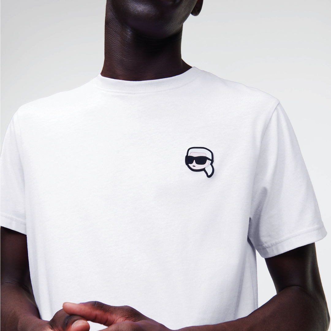 A male model wearing a white crew-neck T-shirt with a black patch on the chest and black trousers, standing against a plain background.