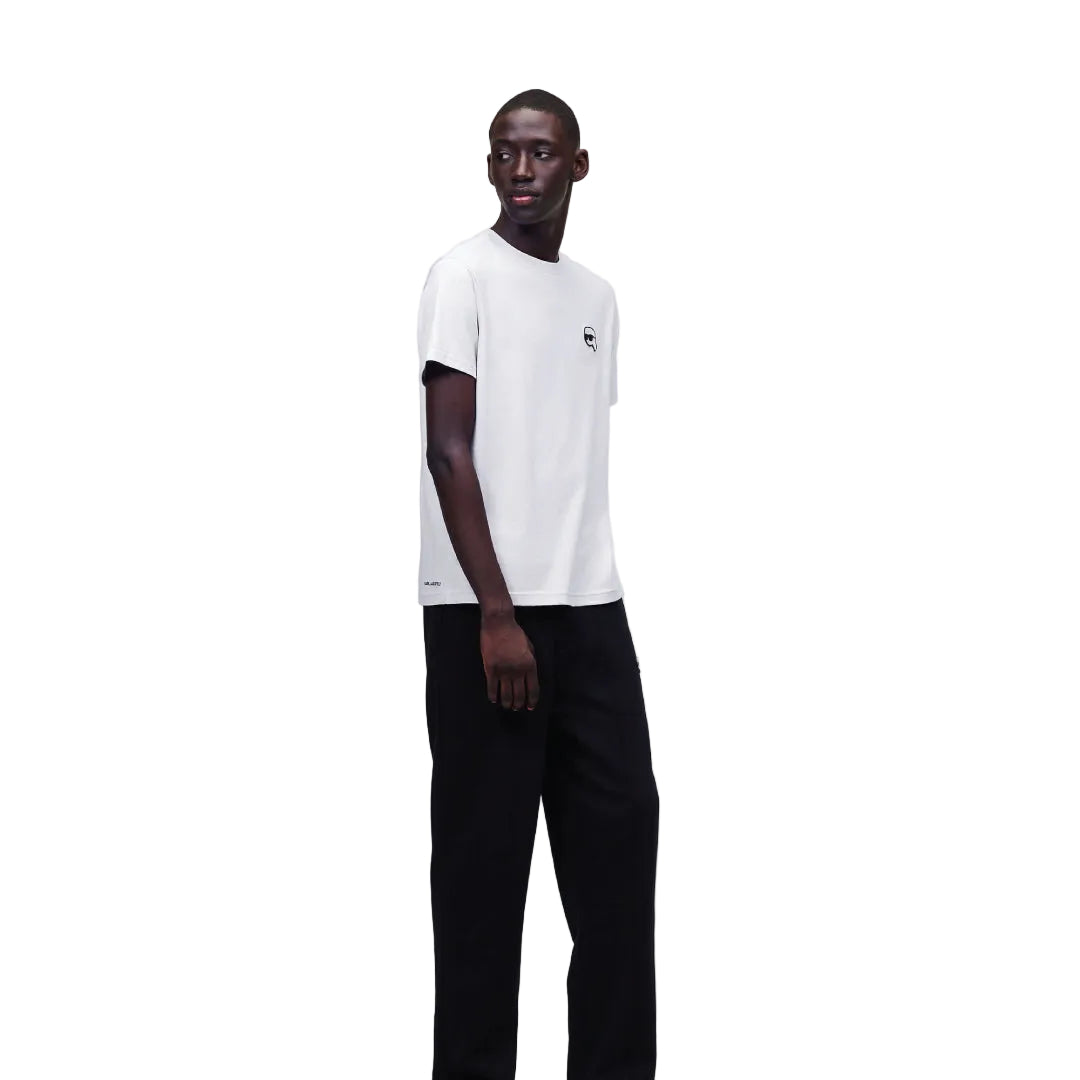 A male model wearing a white crew-neck T-shirt with a black patch on the chest and black trousers, standing against a plain background.