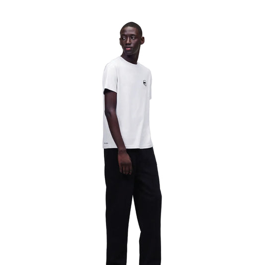 A male model wearing a white crew-neck T-shirt with a black patch on the chest and black trousers, standing against a plain background.