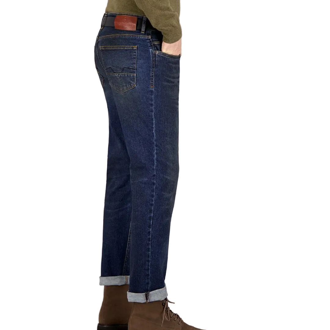 A person standing against a grey background, wearing dark rinse Gardeur jeans with handmade finishings and a rolled-up hem.